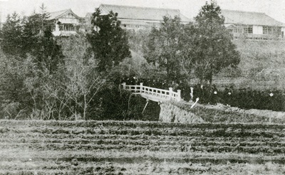 1929年完成の石の養徳橋ができる前の橋。奧が鑵子山校舎。1921年頃。（『天理教校五十年史』より）